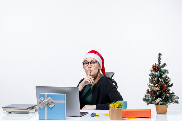 圣诞节戴着圣诞老人帽子和眼镜的严肃的年轻女子坐在一张桌子旁 桌子上放着圣诞树和礼物 在白色背景上做着沉默的手势年轻女人办公室帽子