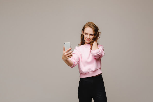 灰色背景一个微笑的快乐女孩穿着粉色衬衫在灰色背景下的智能手机上自拍自拍使用电话