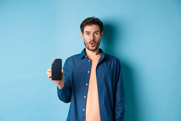 男人印象深刻的白人男模展示智能手机屏幕 看起来很惊讶 谈论网上报价 站在蓝色背景上发型屏幕男性