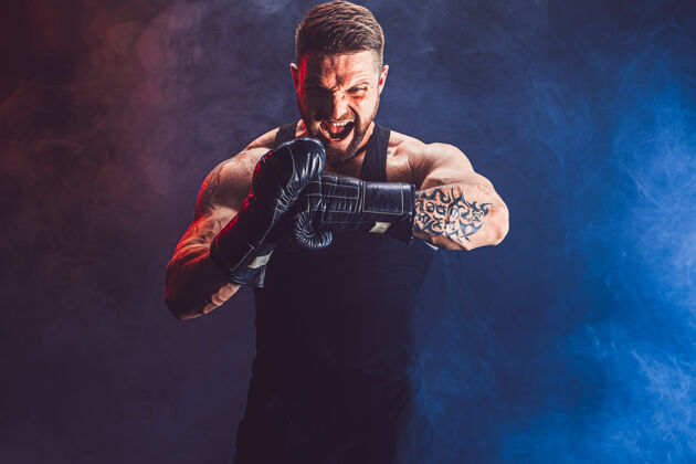 健身留着胡须 纹身的泰拳运动员 身穿黑色汗衫 戴着拳击手套 在黑暗的墙上与烟雾搏斗运动概念肌肉运动员跆拳道