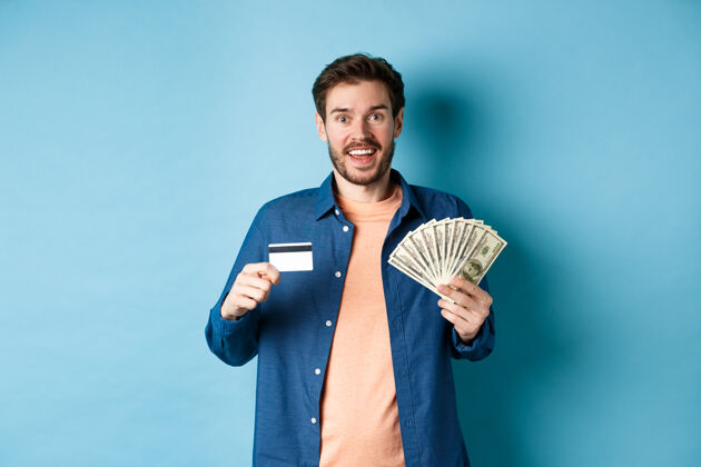 信用卡微笑的白人男子展示塑料信用卡和钱 看起来很高兴 站在蓝色的背景上男人休闲朋友