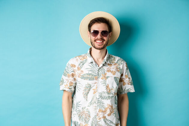 帅气戴着墨镜 穿着夏装 微笑着的帅哥 在旅游中享受假期 站在蓝色的背景上站衬衫帽子