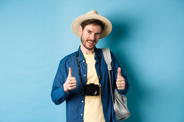 推荐帅气的高加索游客 头戴草帽 背着背包 拿着相机 竖起大拇指 推荐旅行社或度假酒店 蓝色背景相机背包成人