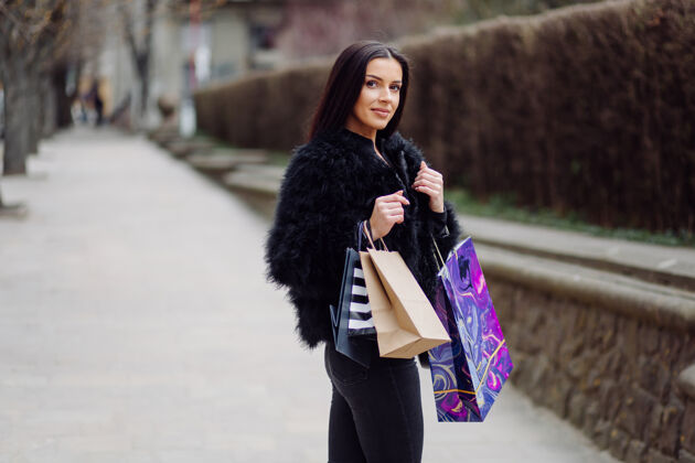 消费者一个棕色头发的女人穿着黑色的衣服 手里拿着五颜六色的有图案的购物袋 在一次成功的购物狂欢中走在外面 她享受着一天的温暖购物购买顾客