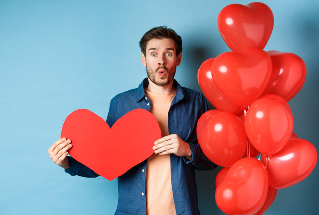 男性情人节的概念恋爱中的男朋友看着惊艳的镜头 捧着红色的纸心 站着浪漫的气球 蓝色的背景伴侣同性恋温柔