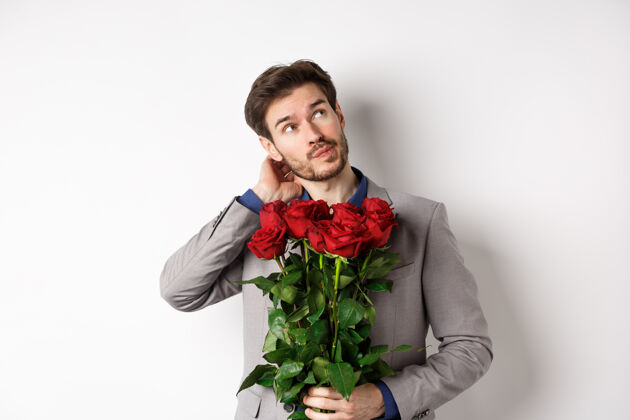 男性体贴的年轻人穿着西装去情人节约会 手里拿着一束玫瑰花 思考着 挠头着 站在白色的背景下微笑情人帅气