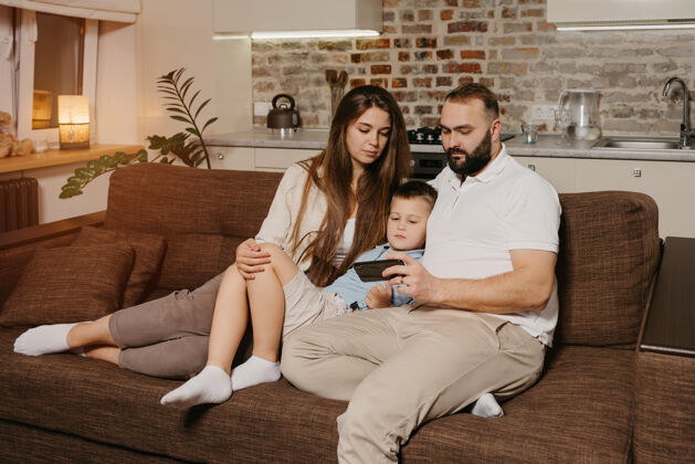 男孩一个爸爸 一个儿子和一个年轻的妈妈在沙发上用智能手机看视频沙发家庭父母身份