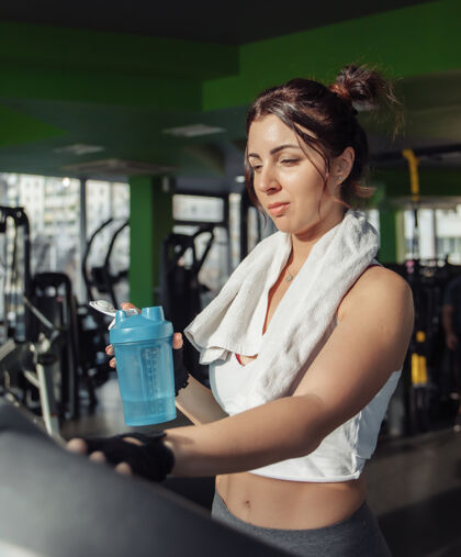 训练一个年轻健康的女人 肩上扛着毛巾 在跑步机上拿着水瓶减肥理念 有氧运动女孩健身房跑步机
