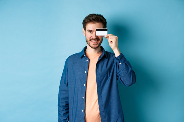 购物者快乐的男客户眼睛上拿着塑料信用卡 微笑着 穿着蓝色背景的休闲装站着自信帅哥男人
