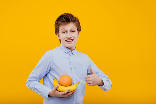年轻可爱的小男孩手里拿着水果 香蕉和橘子 穿着蓝色衬衫 隔离在黄色的墙上秀竖起大拇指英俊