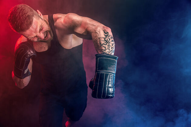 踢留着胡须 纹身的泰拳运动员 身穿黑色汗衫 戴着拳击手套 在黑暗的墙上与烟雾搏斗运动概念年轻私人教练拳击手