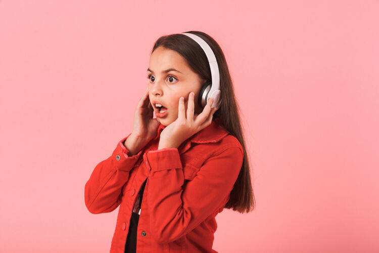 聪明美丽震惊的小女孩用耳机隔着粉色的墙壁听音乐的画面表情耳机花哨