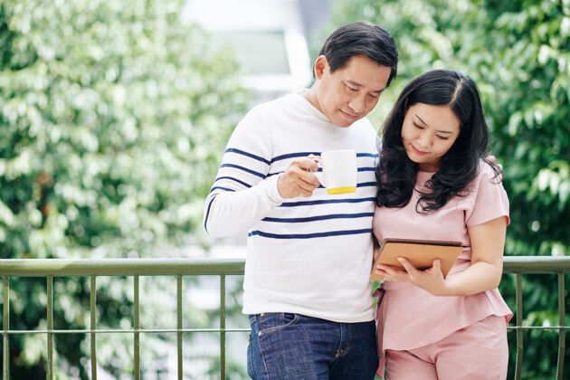休闲活动拥抱站在阳台上的成熟亚洲夫妻 喝着咖啡 在平板电脑上看新闻微笑爱情休闲服