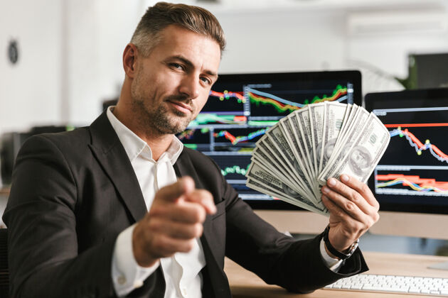 现金令人高兴的30多岁的商人穿着西装拿着钱扇在办公室工作 电脑上有图形和图表肖像财务分析