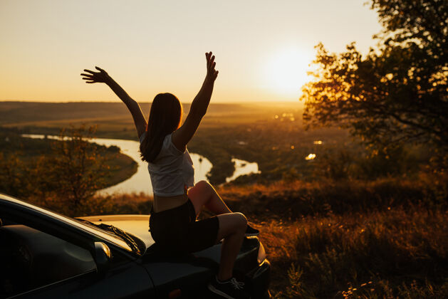 自然坐在车上的年轻女游客举手看美丽的日落的侧视图户外女性美丽