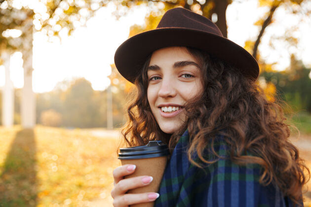 温暖美丽的年轻女子戴着帽子在秋天的公园散步 手里拿着一杯外卖咖啡树叶帽子微笑