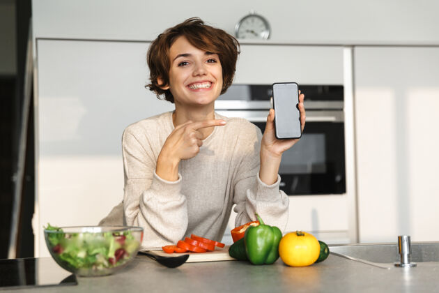 女士快乐的黑发女人在厨房的桌子旁做饭时 展示着空白的智能手机屏幕 并指着屏幕室内吸引看