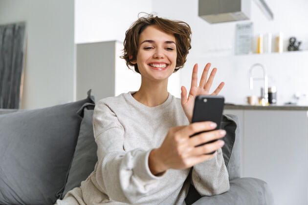 年轻人快乐的年轻女子坐在家里的沙发上 用手机 打视频电话家庭主妇室内应用程序