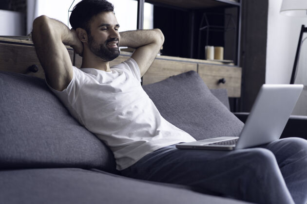 互联网正在键入新的博客文章帅哥坐在家里的沙发上微笑着使用笔记本电脑的侧视图优雅休闲房子