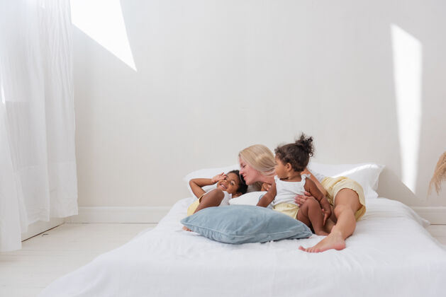 孩子年轻的白人金发母亲玩耍 和非裔美国人的女儿儿子躺在白色的床上女人女儿女性