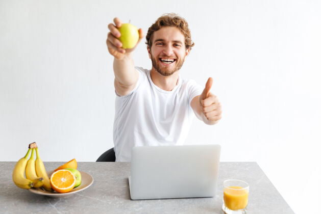 学习照片中 一个快乐的留着胡子的年轻人在家里的桌子旁用笔记本电脑拿着水果 摆出竖起大拇指的姿势吸引水果年轻