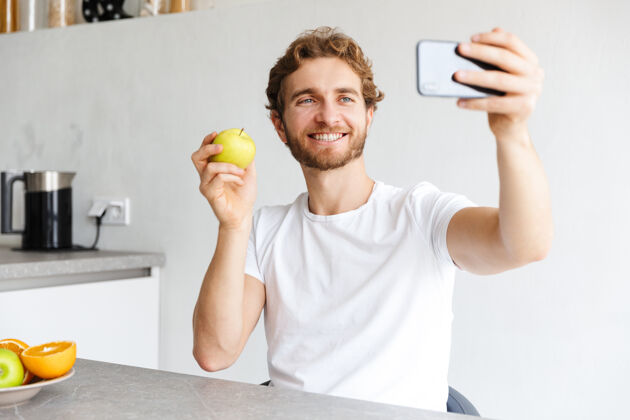 软图为一位快乐的留着胡子的年轻人在家里的餐桌上用水果通过电话自拍室内放松白种人