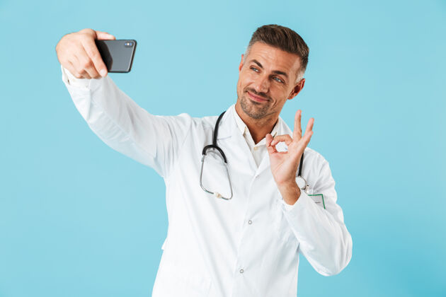 阳性照片中的医生身穿白大褂 手持听诊器在智能手机上自拍 孤零零地站在蓝色的墙上中年成熟手机