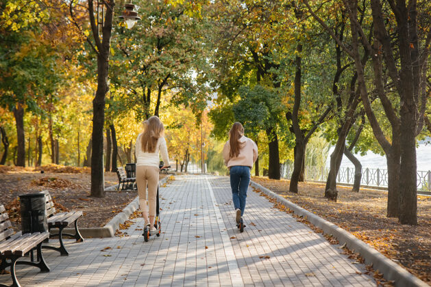 背部在温暖的秋天 年轻漂亮的姑娘们骑着电动滑板车在公园里日间步行在公园里道路科技周末