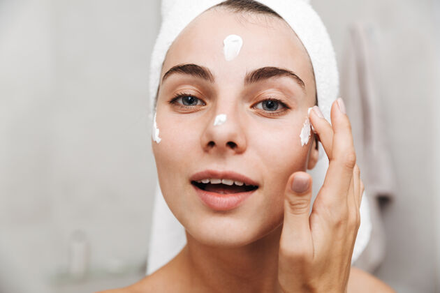 白种人特写镜头：一个头上戴着毛巾的年轻漂亮女人站在浴室里涂面霜卫生面部化妆
