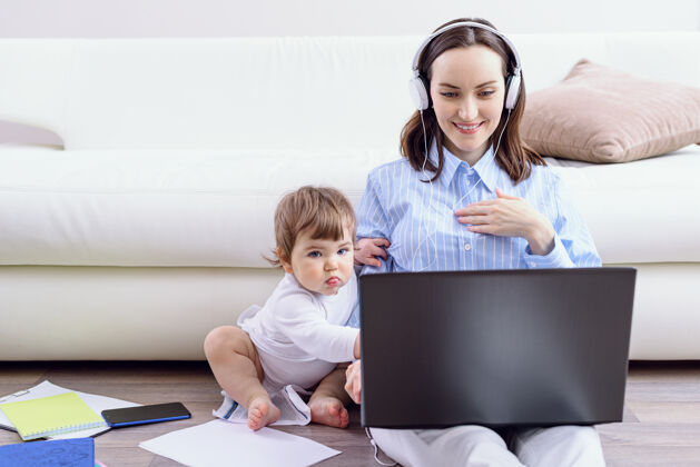 互联网戴着耳机的女人看着笔记本电脑屏幕 孩子正坐在她旁边家庭培训企业家儿童