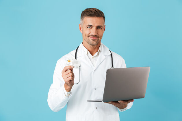 中年照片中的白种人身穿白色医用外套 手持手提电脑和信用卡的听诊器 孤立地站在蓝色的墙上职业医院男人
