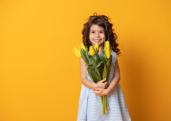 活动穿着条纹连衣裙的可爱小女孩手持一束郁金香在黄色的工作室里背景.spring妇女节酷生日女孩