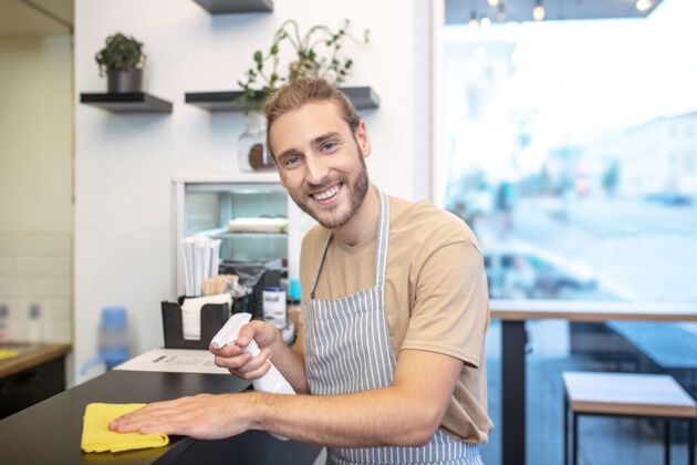 表面伟大的心情微笑年轻的留着胡须的帅哥拿着消毒液和餐巾站在咖啡馆的柜台旁擦拭着表面消毒工作日工作
