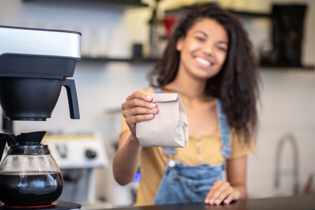 成功咖啡小包装咖啡馆柜台后面站着一位兴高采烈的长发女咖啡师 她伸出手中的纸袋工作日腰向上创业