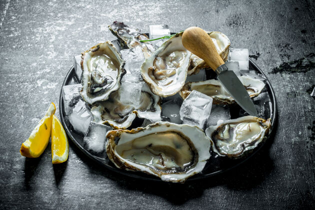 深色新鲜生牡蛎放在盘子里 用冰块 刀和柠檬片放在深色的木桌上海洋餐桌软体动物