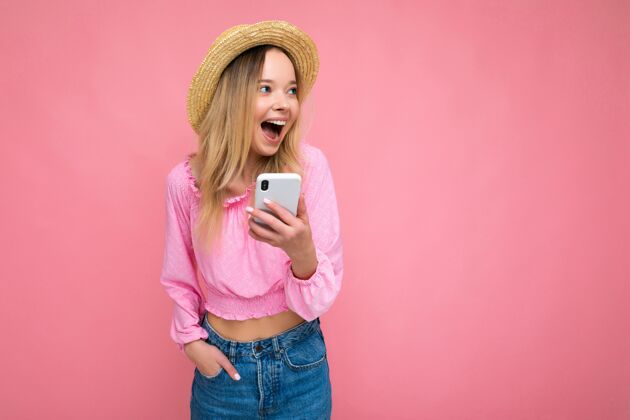 通信迷人迷人的金发女郎微笑着 开心地站在粉色的墙上 手放在口袋里 穿着粉色的衬衫 戴着夏天的帽子 用手机看着旁边轻松微笑技术