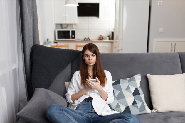坐着手拿手机坐在舒适沙发上的女人休闲周末科技年轻美容房间