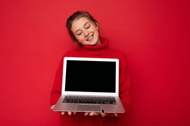 网页照片中 美丽的知足快乐的年轻女子手持电脑笔记本 身穿红色毛衣隔着红墙低头看着上网本屏幕背景.空空间工作空闲时间上网本