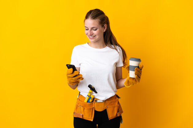 饮料年轻的电工女孤立在黄色的背景下拿着咖啡带走和移动手机采取技能