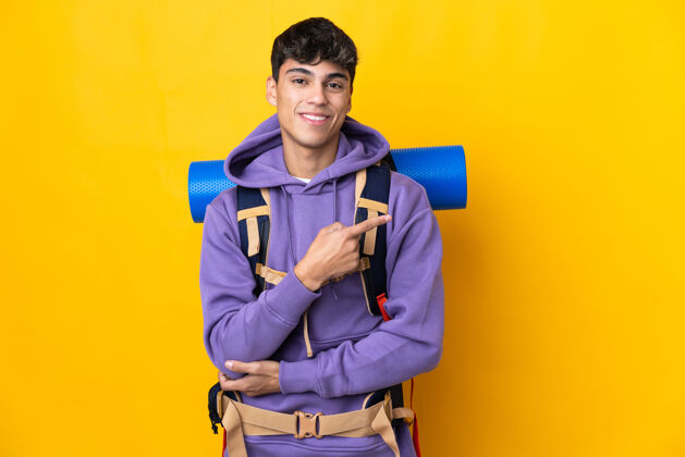 男人年轻的登山男子背着一个大背包 在孤立的黄色背景下指着旁边展示一款产品徒步旅行徒步旅行运动