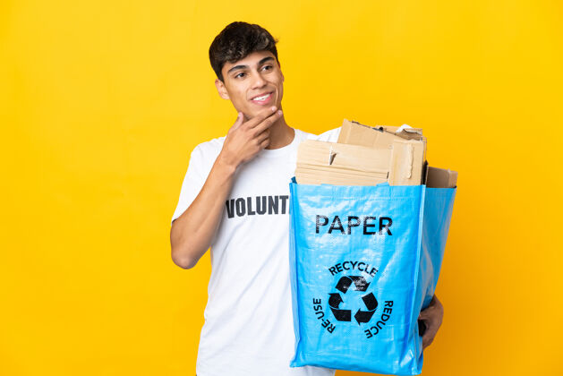 年轻一个男人拿着一个装满废纸的回收袋 站在隔离的黄色墙壁上 微笑着抬起头来幸运垃圾男性