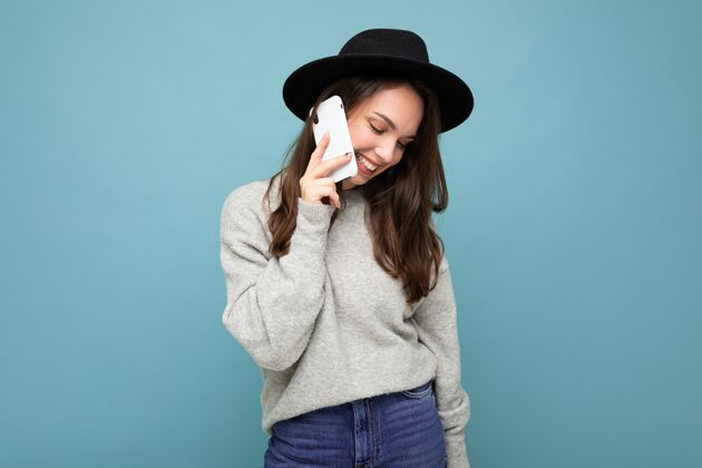 手机迷人的年轻腼腆微笑的女人戴着黑色帽子和灰色毛衣拿着智能手机在背景上孤立地往下看移动嘿好心情