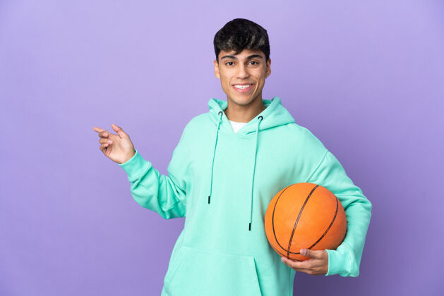 运动员年轻人在孤立的紫色背景上打篮球 手指指向侧面展示快乐健康
