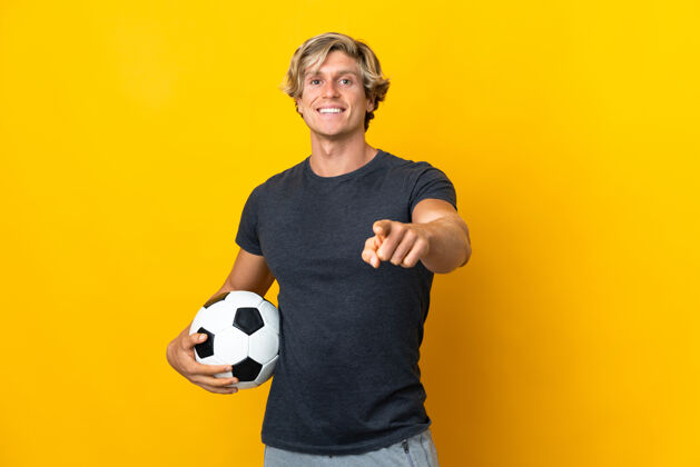 运动员英国人在孤立的黄色背景上用足球指着前面训练休闲帅哥