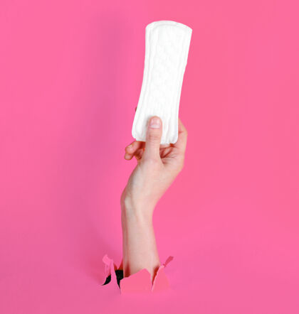 月经女性手拿卫生巾穿过撕破的粉色纸背景：极简主义时尚理念月产品定期