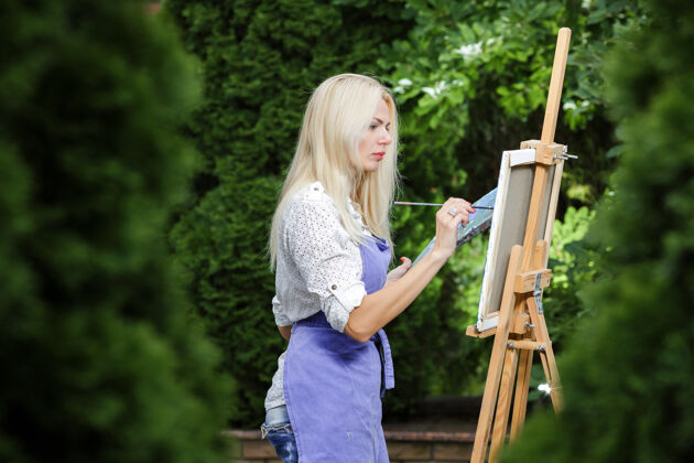 人物美丽的金发女画家手里拿着画笔在花园里的画布上画画成人自然画家