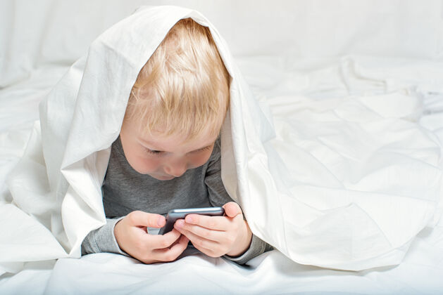 谎言金发小男孩正在电视上看什么智能手机撒谎躺在床上 躲在被窝里童年科技小