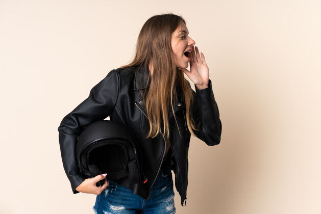 喊叫立陶宛一名年轻女子手持摩托车头盔 孤立地站在米色背景下 张大嘴巴向一侧喊叫驾驶声音情绪