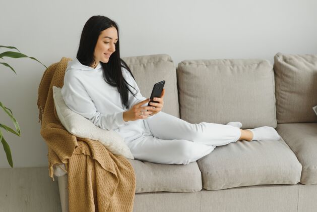 休闲一个快乐的年轻女子在家里的沙发上休息时使用手机的画像坐着抱着放松