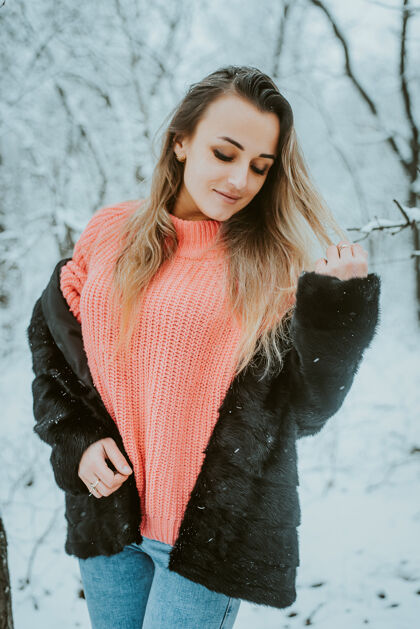 雪在寒冷的雪域森林里 穿着皮草外套 粉色宽松毛衣和牛仔裤的美丽少女公园降雪女孩